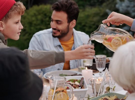 Faceless female pouring fruit beverage into glass of boy in warm cloth while cheerful multiethnic family having dinner outdoors
