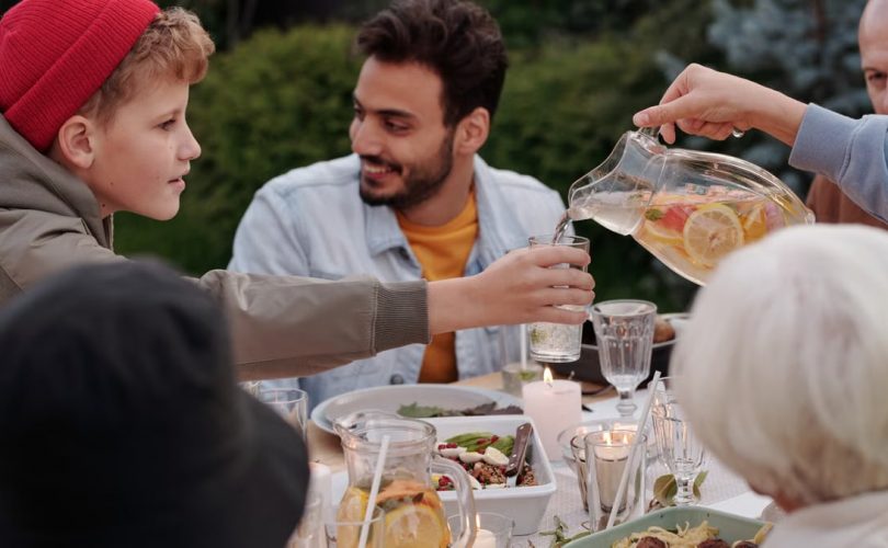 Faceless female pouring fruit beverage into glass of boy in warm cloth while cheerful multiethnic family having dinner outdoors