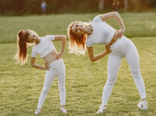 A mother and daughter in activewear stretching on a sunny day in a grassy field.