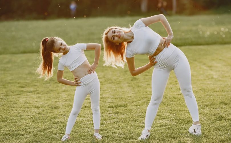 A mother and daughter in activewear stretching on a sunny day in a grassy field.