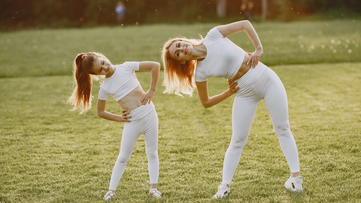 A mother and daughter in activewear stretching on a sunny day in a grassy field.